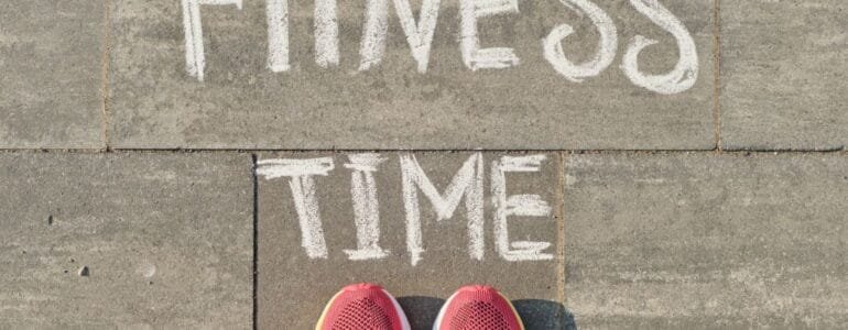 Text fitness time written on gray pavement with woman legs in sneakers, view from above